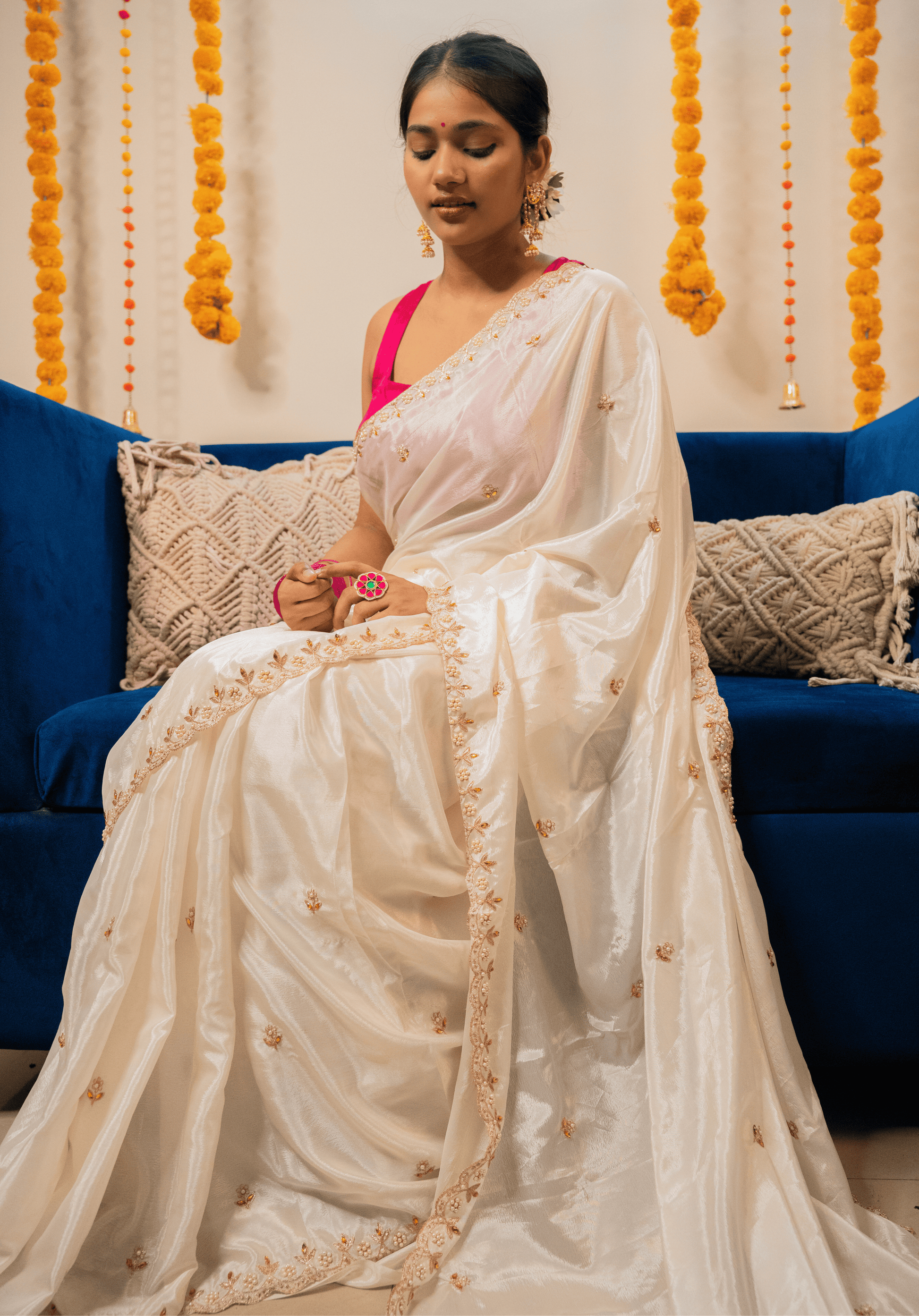 Woman in a white chinon saree with gold details sitting on a blue couch with decorative elements in the background.