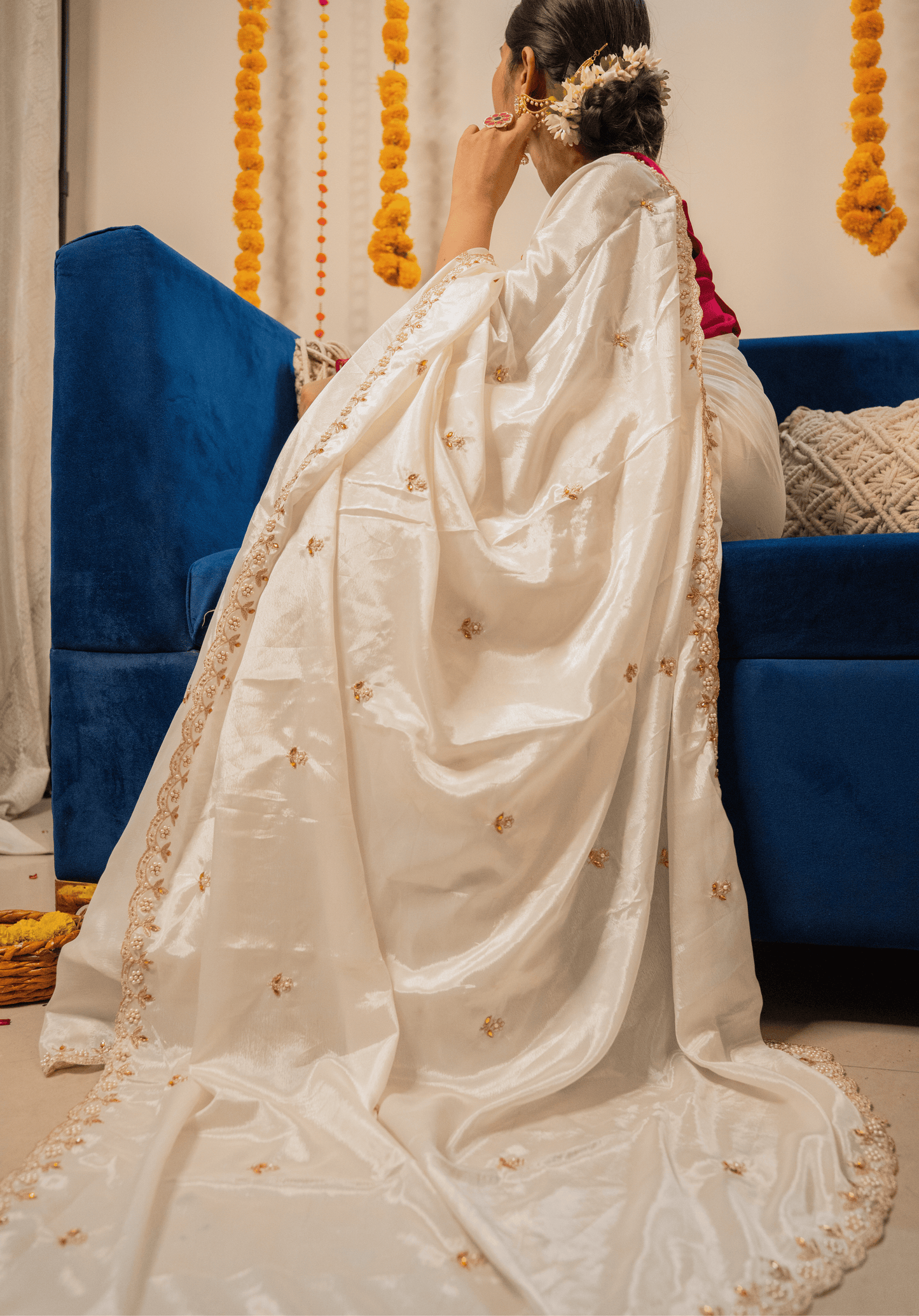Person wearing a white saree with gold details in a decorated room.