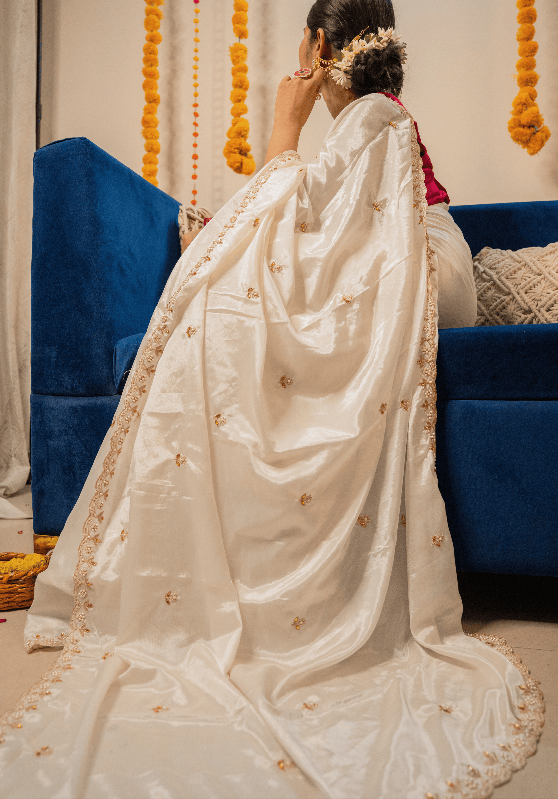 Person wearing a white saree with gold details in a decorated room.