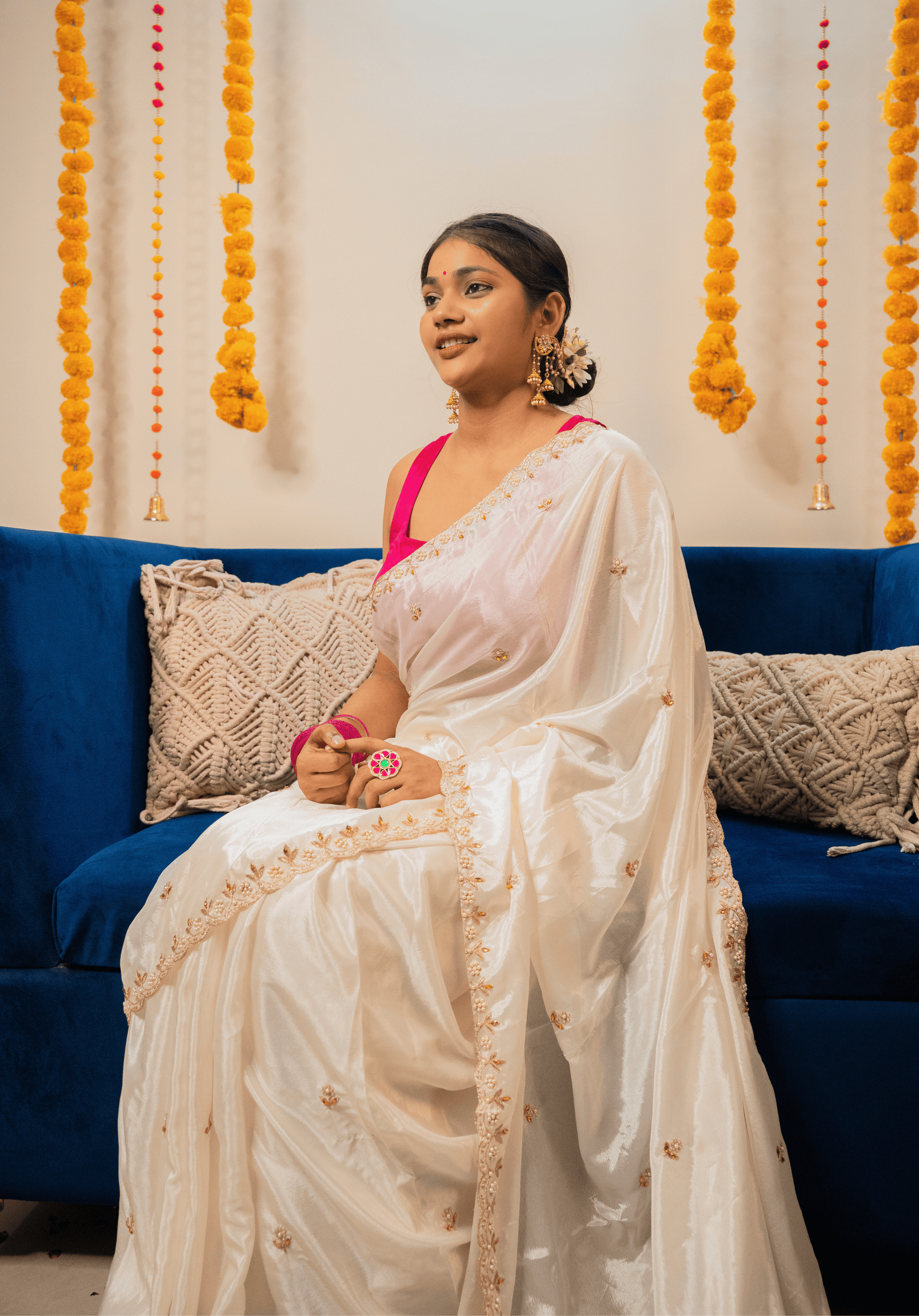 Woman in a white chinon saree with a pink blouse sitting on a blue couch with decorative elements in the background.