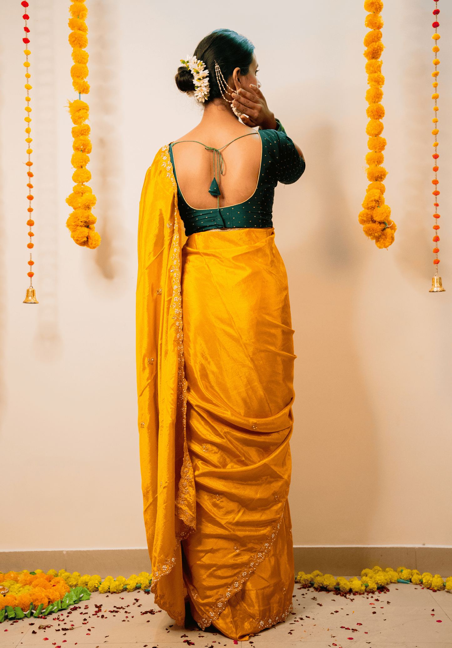 Woman in a yellow chino saree with a green blouse standing against a decorated wall.