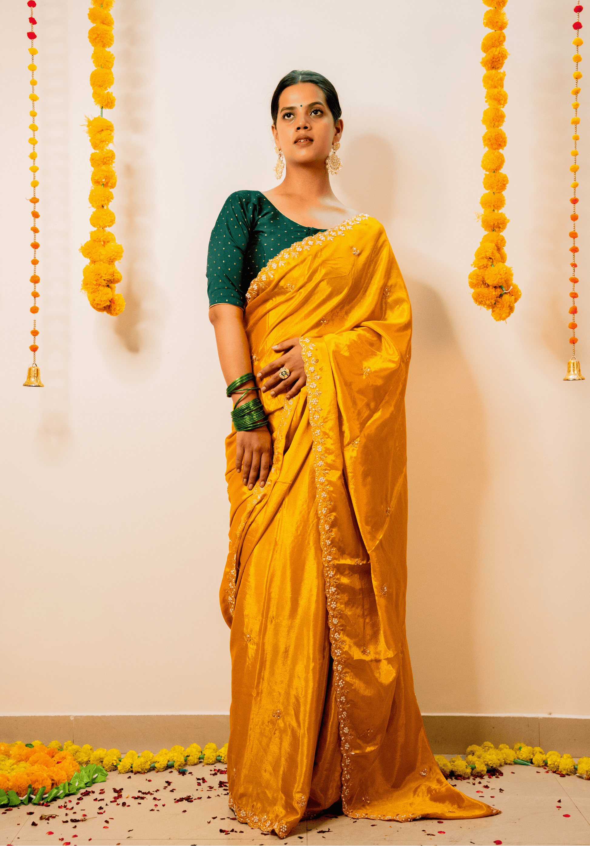Woman in a yellow saree with green blouse standing against a decorated wall.