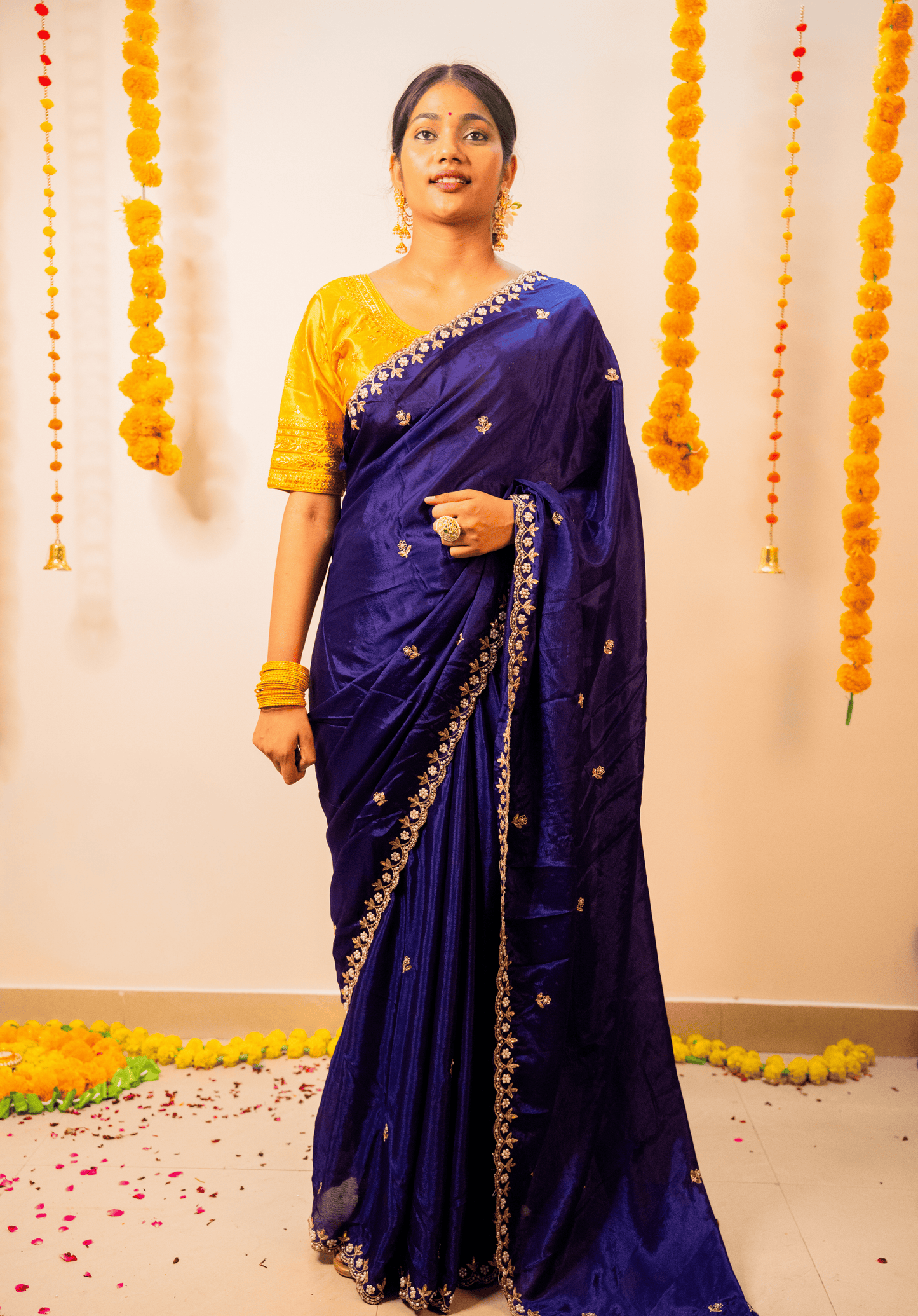 Woman in a purple chinon saree with a yellow blouse standing against a decorated wall.