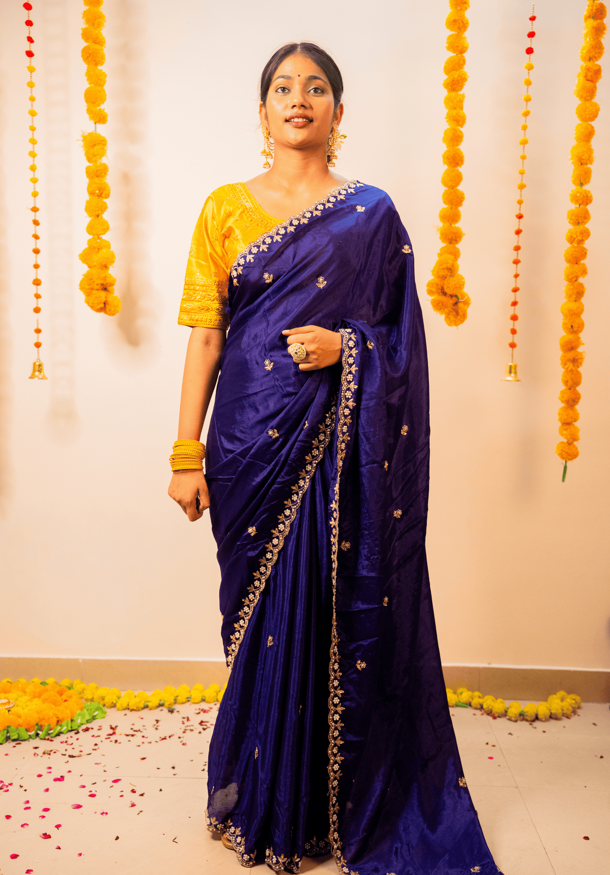 Woman in a purple chinon saree with a yellow blouse standing against a decorated wall.