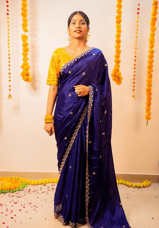 Woman in a purple chinon saree with a yellow blouse standing against a decorated wall.
