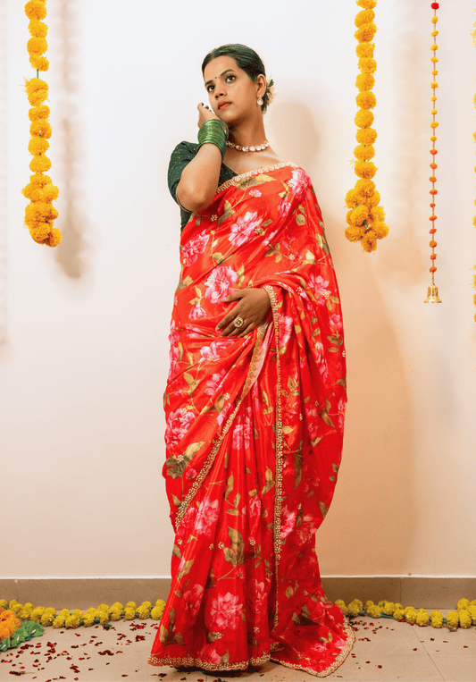 Woman wearing a cherry red floral chinon saree with a green blouse against a decorated wall.