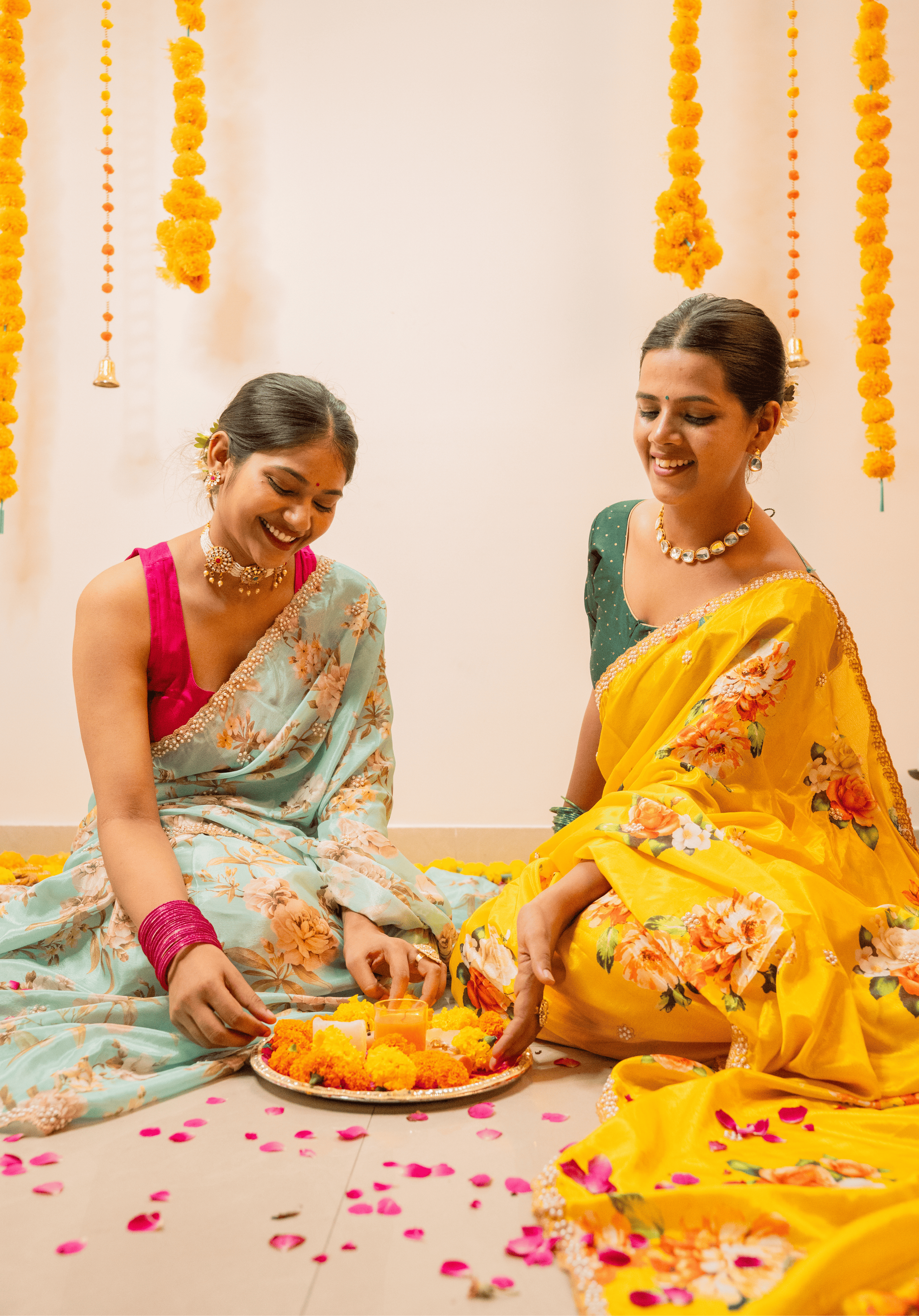 Two women in traditional sarees sitting on a decorated floor with flowers and plates.