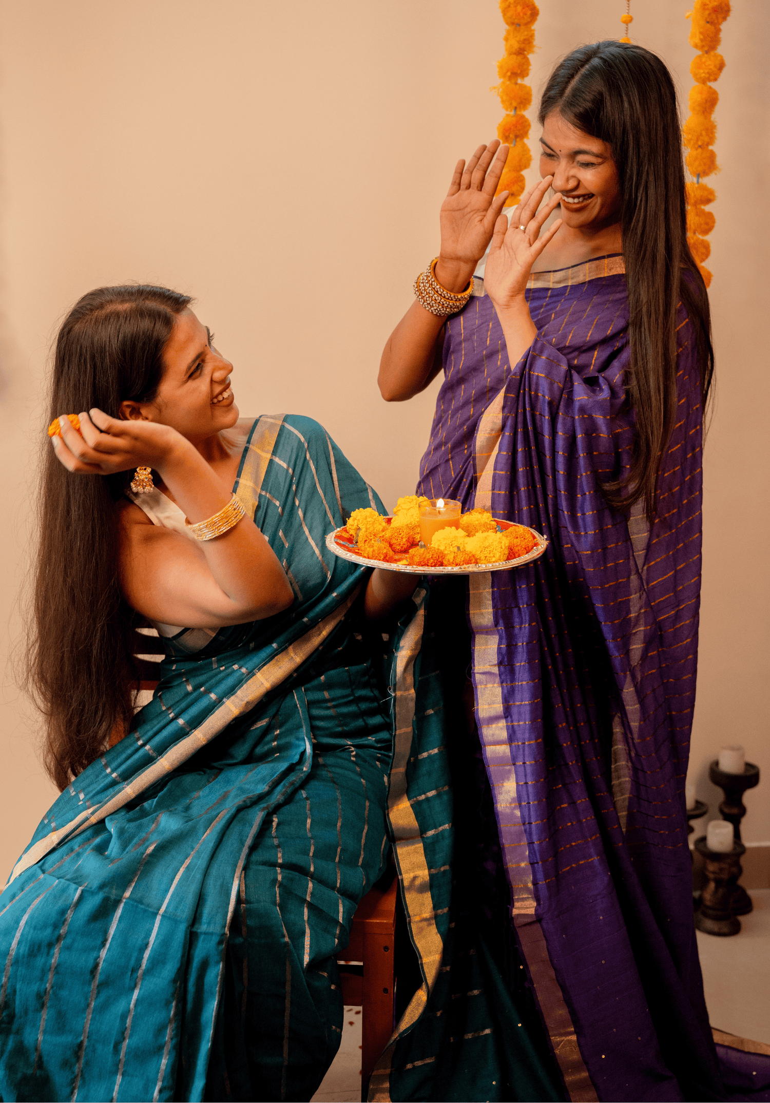 Two women in traditional sarees with one holding a plate of flowers, against a neutral background.
