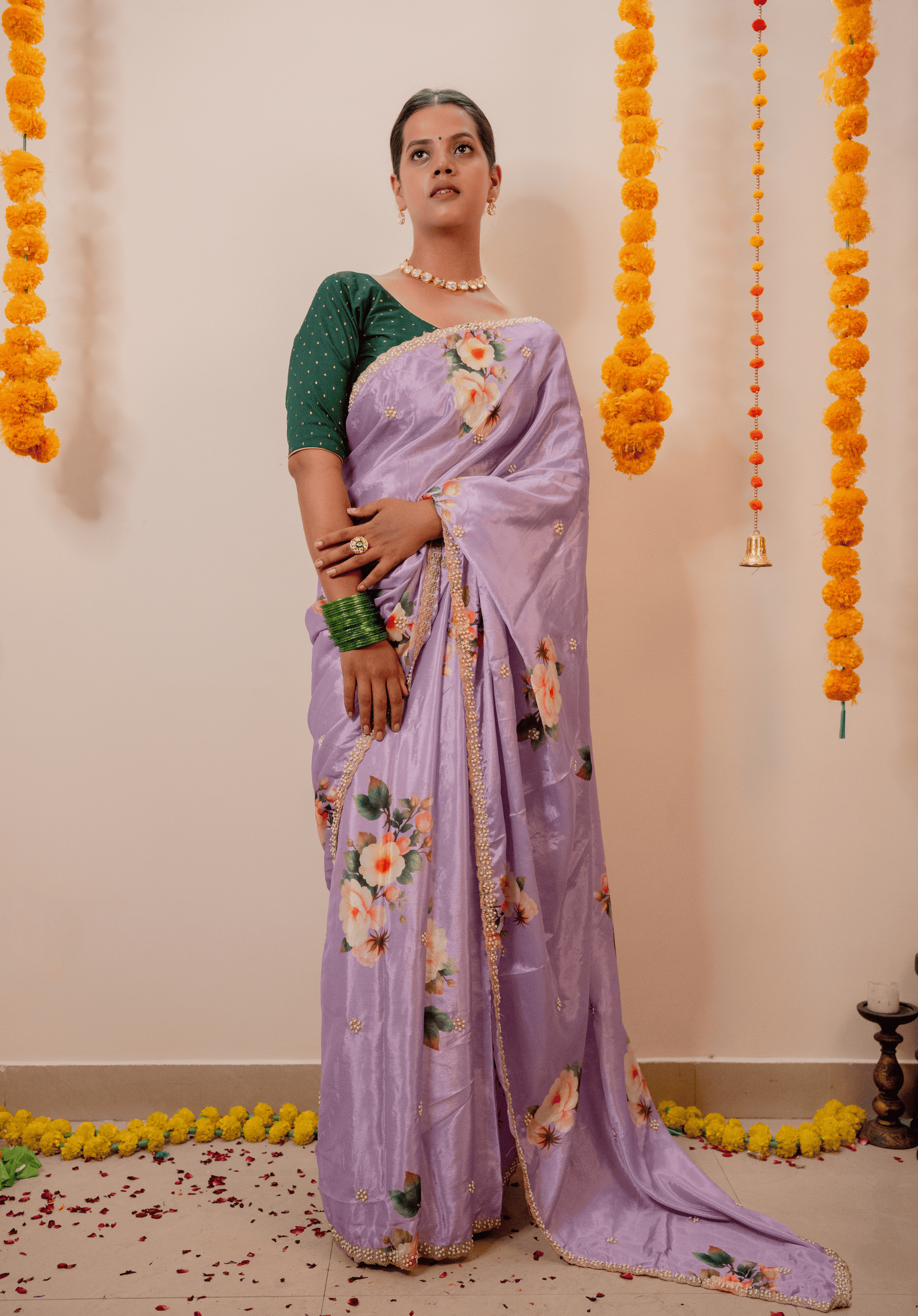 Woman in a lilac chinon saree with floral patterns standing against a decorated wall with marigold flowers.
