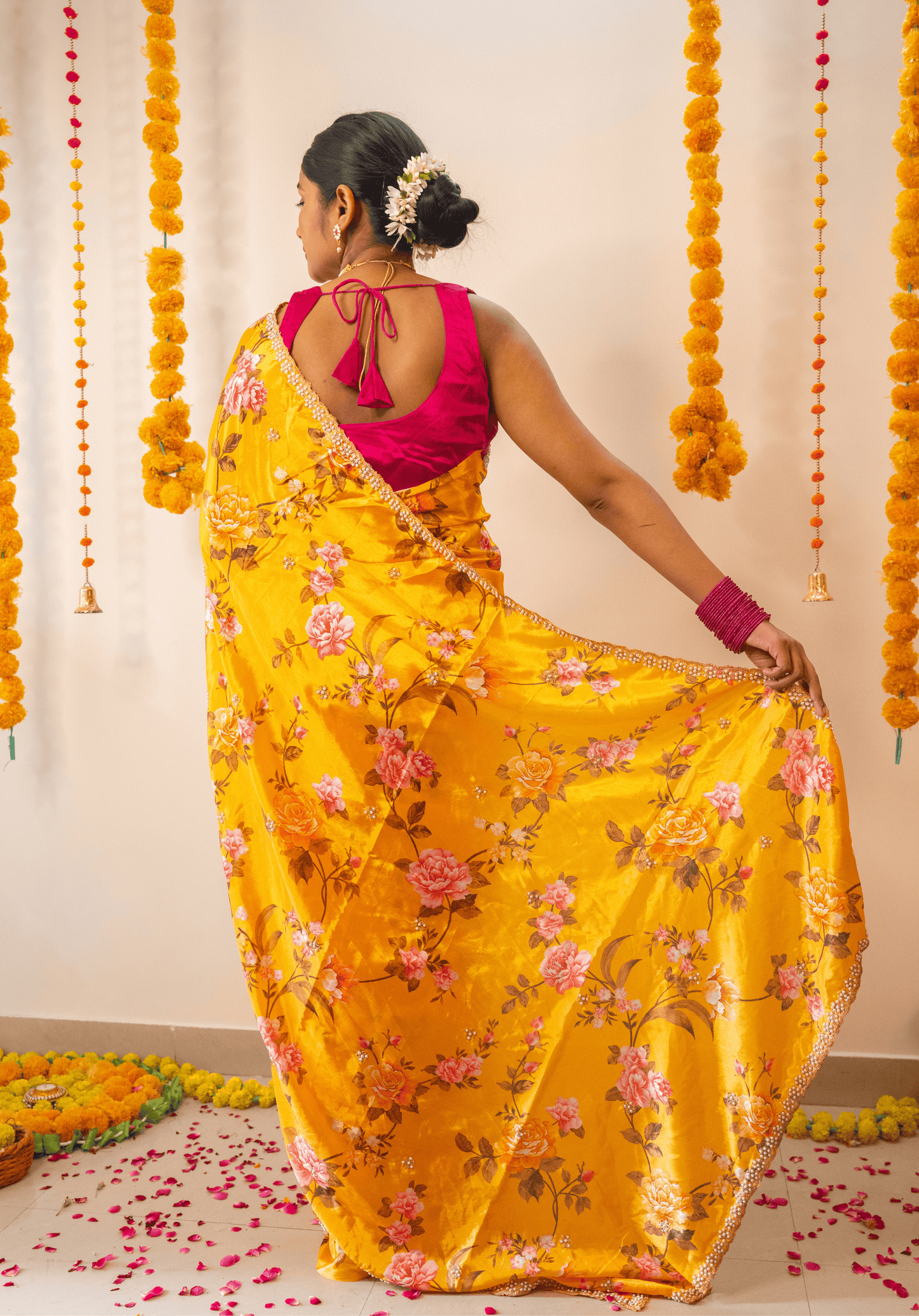 Woman in a mustard yellow floral embroidered chinon saree with a pink blouse standing in front of decorative marigold flowers.