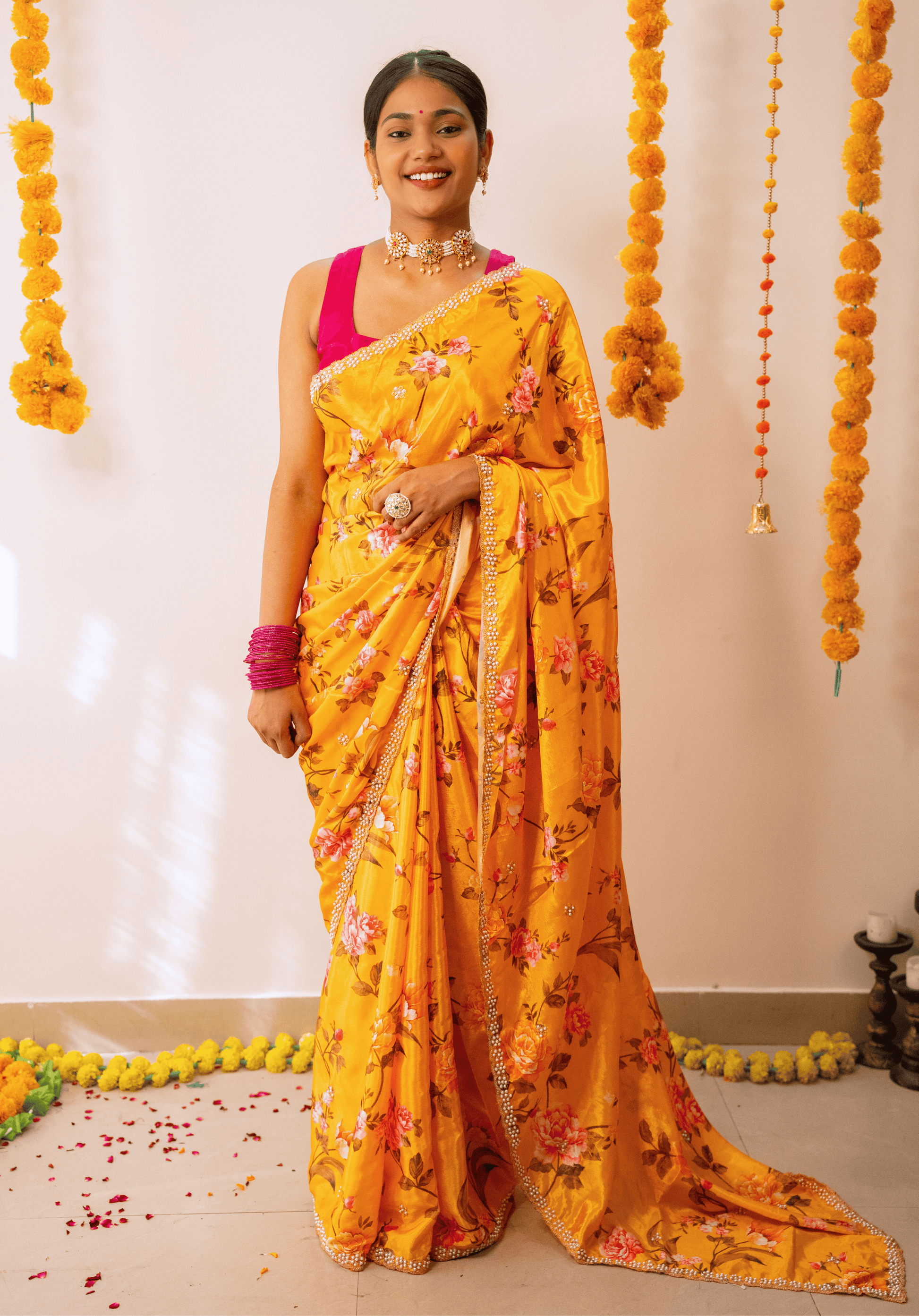 Woman in a mustard yellow floral embroidered chinon saree with pink blouse standing against a decorated wall.