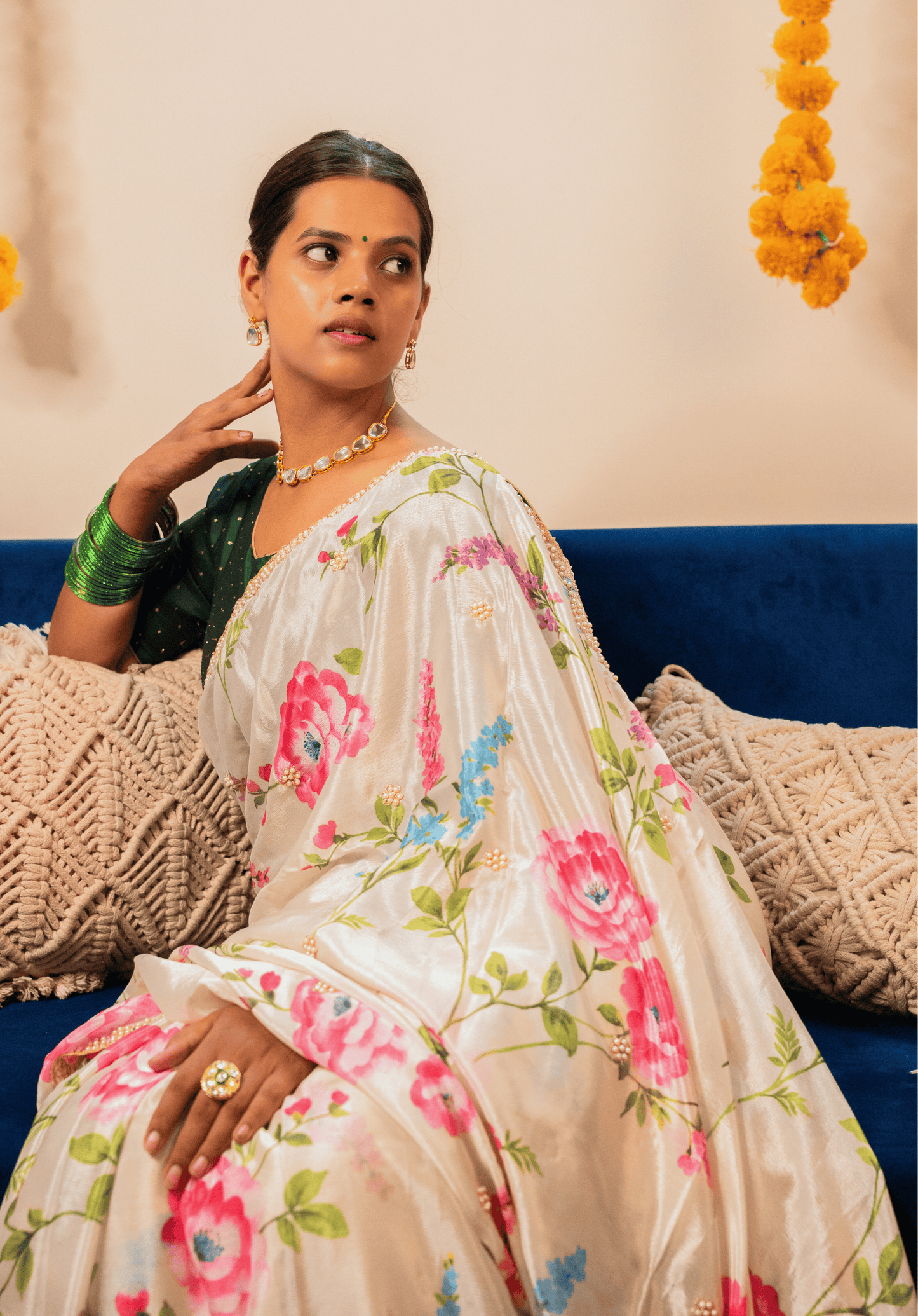 Woman in an off white floral chinon saree sitting on a blue couch with decorative elements in the background