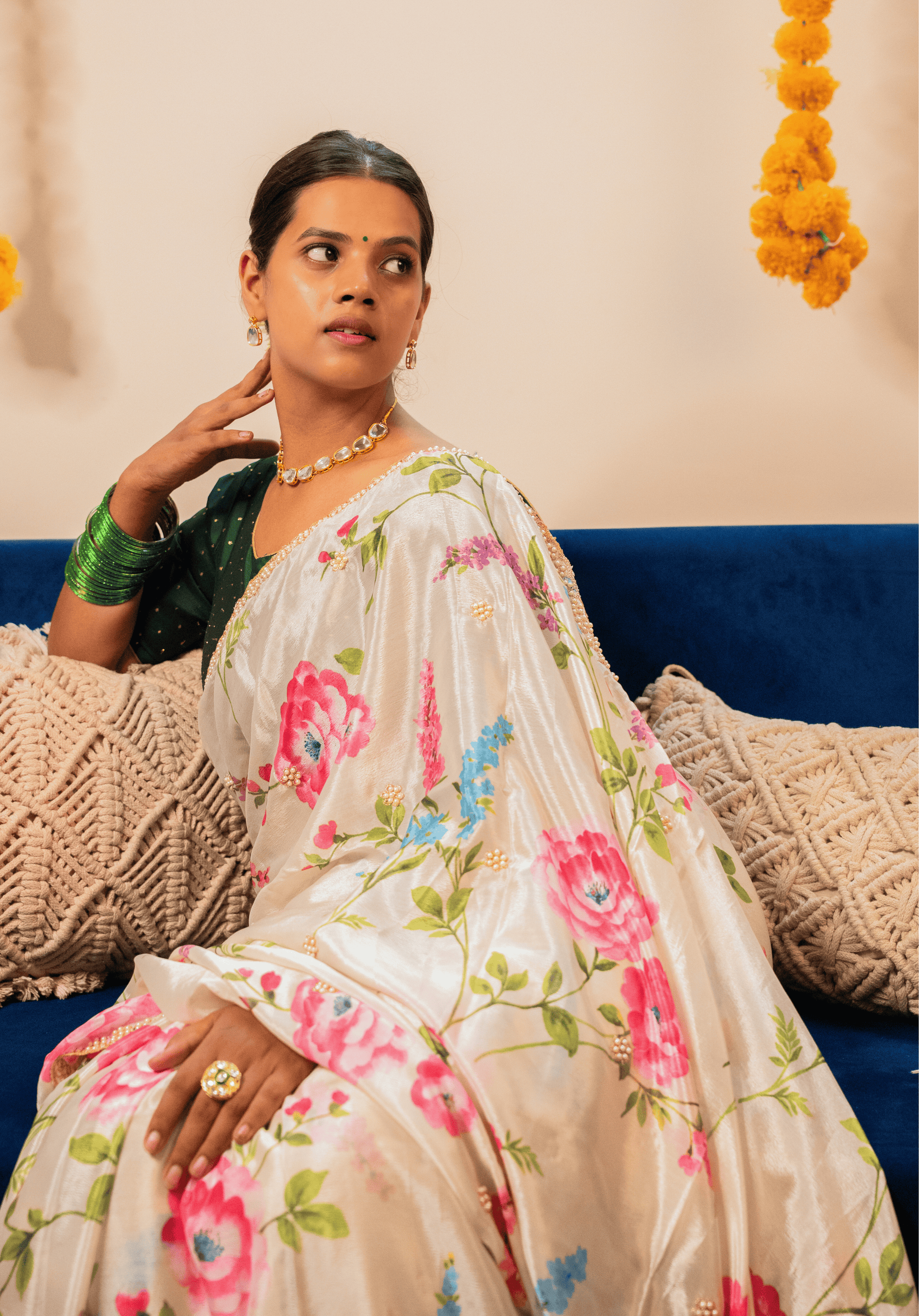 Woman in an off white floral chinon saree sitting on a blue couch with decorative elements in the background