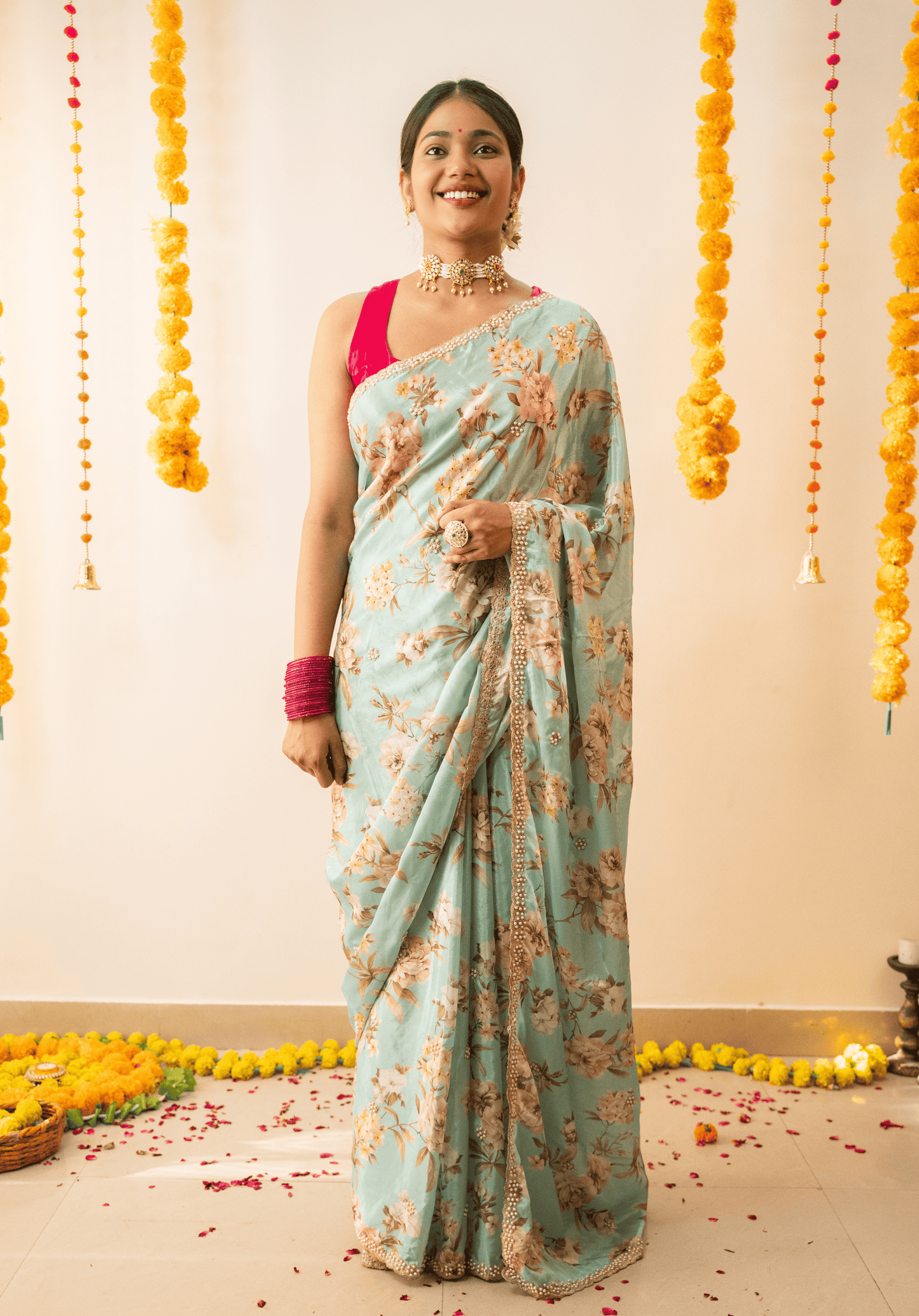 Woman in a pastel green floral chinon saree with a pink blouse standing against a decorated wall.