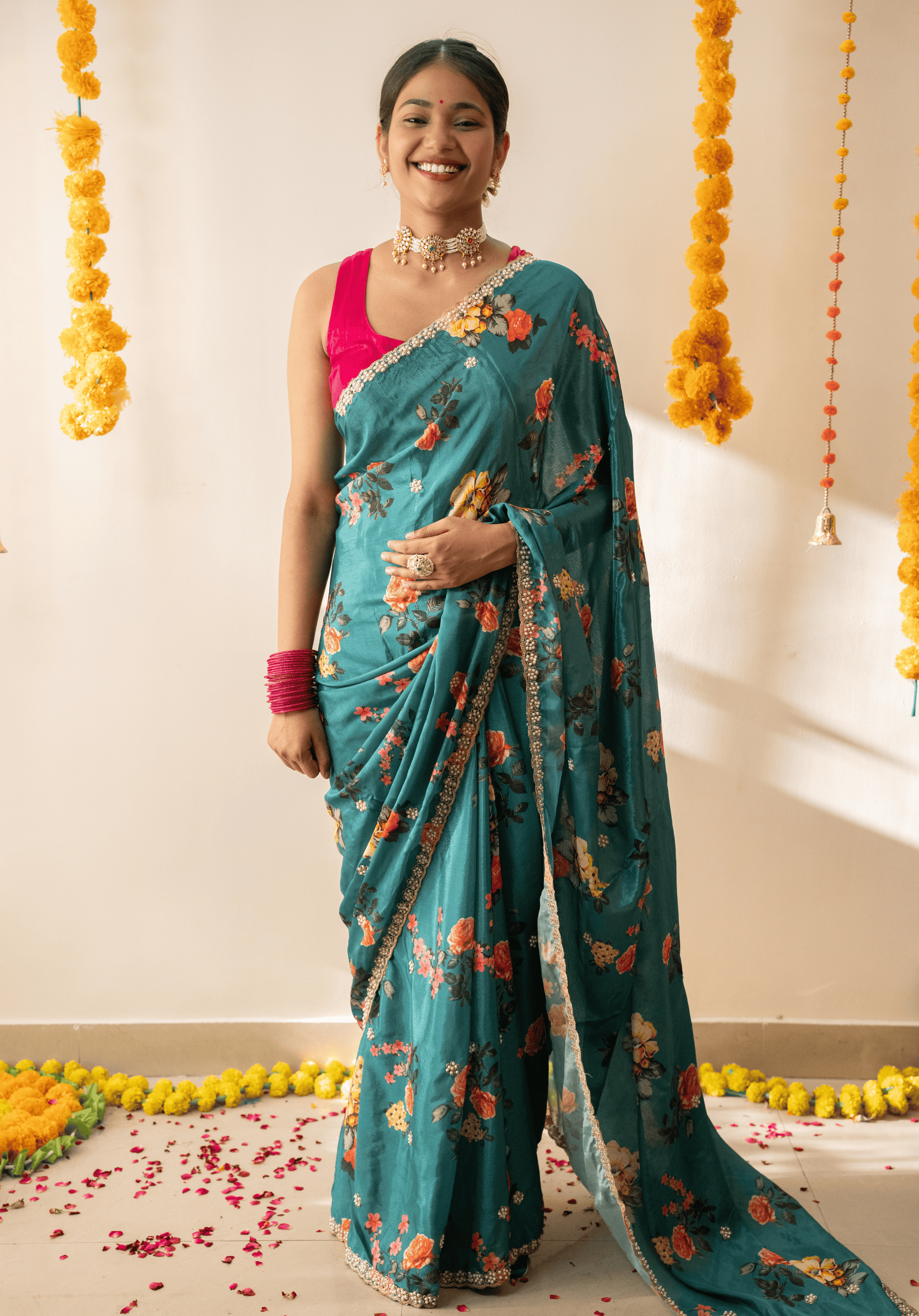 Woman in a teal green chinon saree with floral patterns and pink blouse standing against a decorated background.
