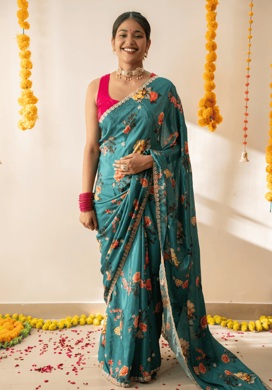 Woman in a teal green chinon saree with floral patterns and pink blouse standing against a decorated background.